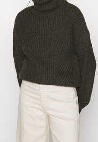 Pullover di lana chunky verde scuro con un alto collo a patella e texture a costine. Abbinato a pantaloni larghi di colore crema chiaro.