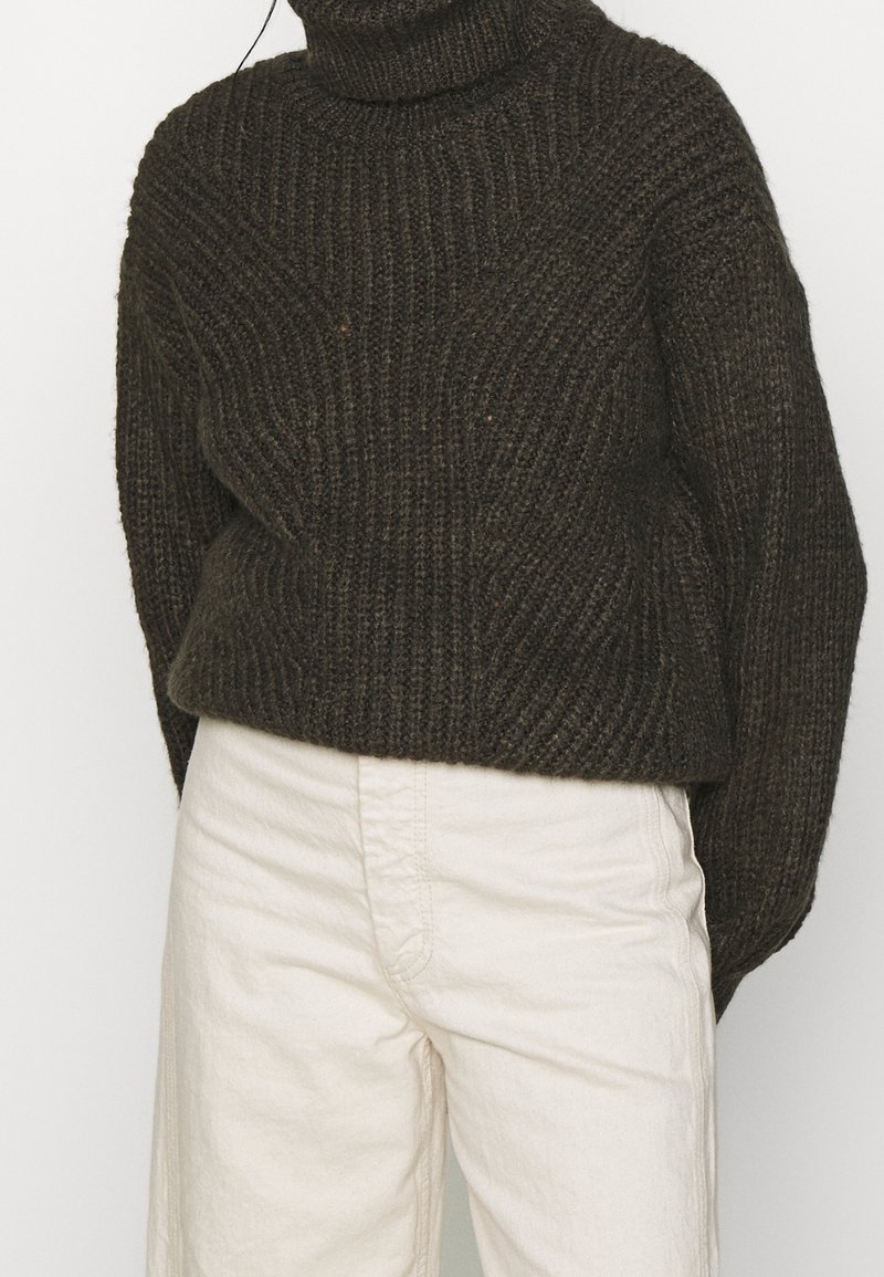Pullover di lana chunky verde scuro con un alto collo a patella e texture a costine. Abbinato a pantaloni larghi di colore crema chiaro.