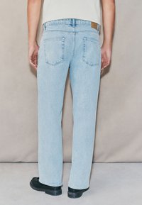 Light blue denim jeans with a straight leg cut, two back pockets, and a tan leather patch at the waistband. Black shoes are worn.