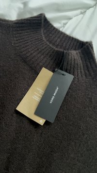 Mørkebrun ribbestrikket genser med rund hals, med svart merkelapp og en tan fargepleieetikett festet. Laget av polyester.