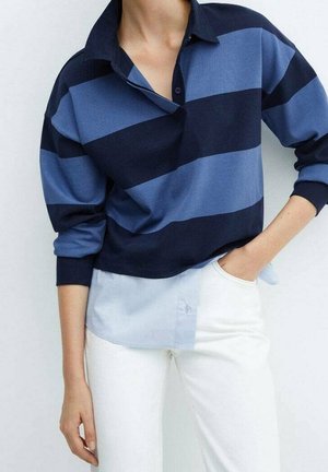 Personne portant un haut à manches longues rayé horizontalement bleu marine et bleu clair sur une chemise bleu clair boutonnée, associée à un pantalon blanc.