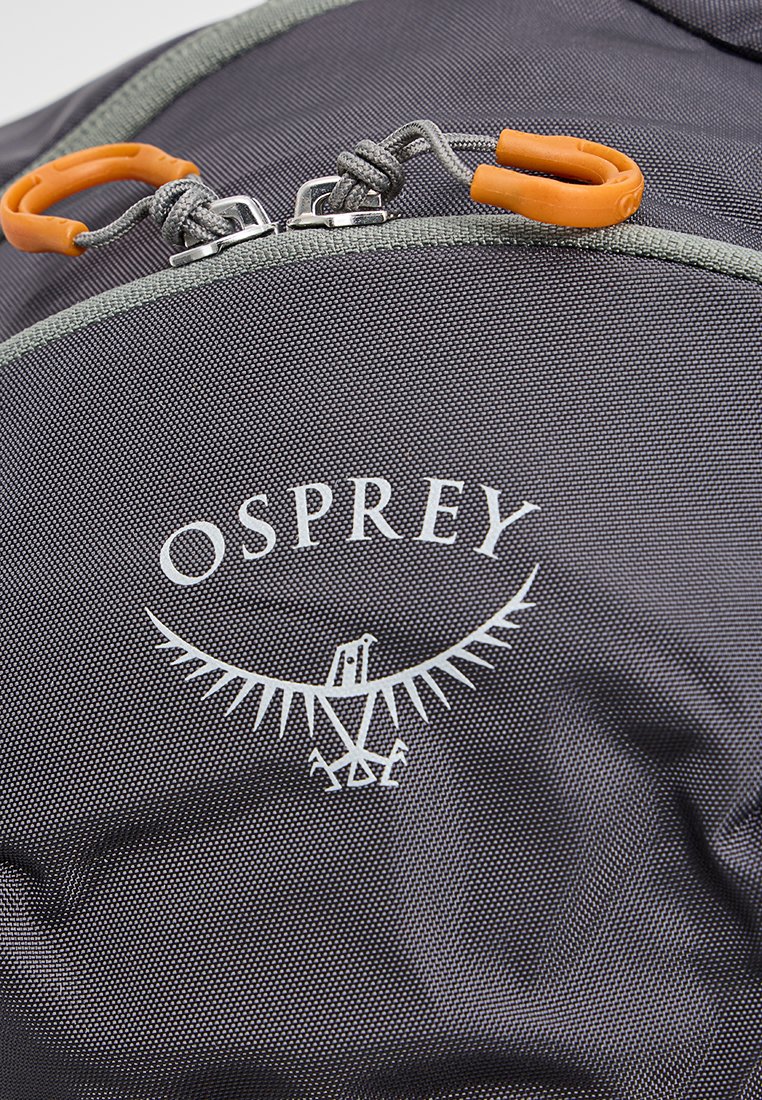 Mochila de tela negra con un logotipo plateado de un águila y cremalleras con tiradores naranjas. La textura es fina y duradera, diseñada para la funcionalidad.