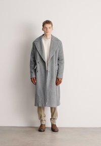 Γκρι μάλλινο παλτό με φαρδιούς γιακάδες, διαθέτει διπλό κούμπωμα και πλαϊνές τσέπες, φορεμένο πάνω από ένα ανοιχτόχρωμο ριπ πλεκτό πουλόβερ και μπεζ παντελόνι.