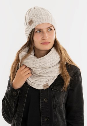 Lichtbeige gebreide beanie met ribbeltextuur en klein logo; bijpassende oneindigheidssjaal in hetzelfde materiaal en kleur, gedragen over een zwarte jas.