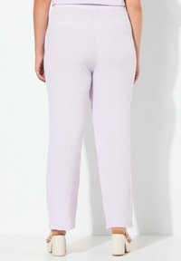 Vrouw in lichtpaarse, gestructureerde broek en witte block-heeled sandalen, van achteren weergegeven tegen een witte achtergrond.