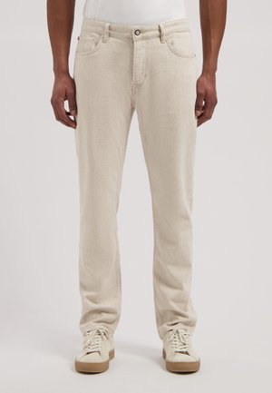 Man draagt lichtbeige recht gesneden spijkerbroek en gebroken witte sneakers, staand tegen een egale achtergrond.