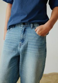 Blå denimjeans med løs pasform, lys vask, synlige syninger og sidelommer, kombineret med en mørkeblå kortærmet skjorte.
