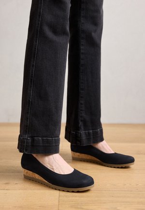 Schwarze Wedgeschuh mit glattem Obermaterial und Korkakzenten am Absatz. Die Sohle hat ein Zickzackmuster für besseren Halt. Getragen mit schwarzen Jeans.