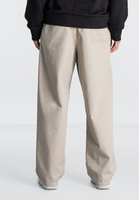 Beige vide bukser med teksturert stoff, med høy midje og en ren kant, kombinert med en svart sweatshirt og grå joggesko.