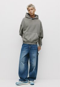 Gray oversized hoodie with a front pocket, paired with loose blue denim jeans featuring distressed details. Shoes are blue and white layered design.