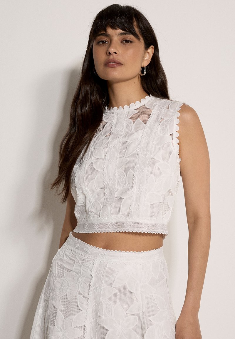 Frau mit langen dunklen Haaren, die ein ärmelloses weißes Spitzen-Crop-Top und einen dazu passenden hochtaillierten Rock mit Blumenmustern trägt.