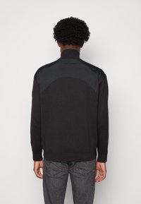 Maglione nero con colletto alto, caratterizzato da un corpo a maglia con texture e pannelli delle spalle lisci in contrasto. Polsini e orlo a costine.