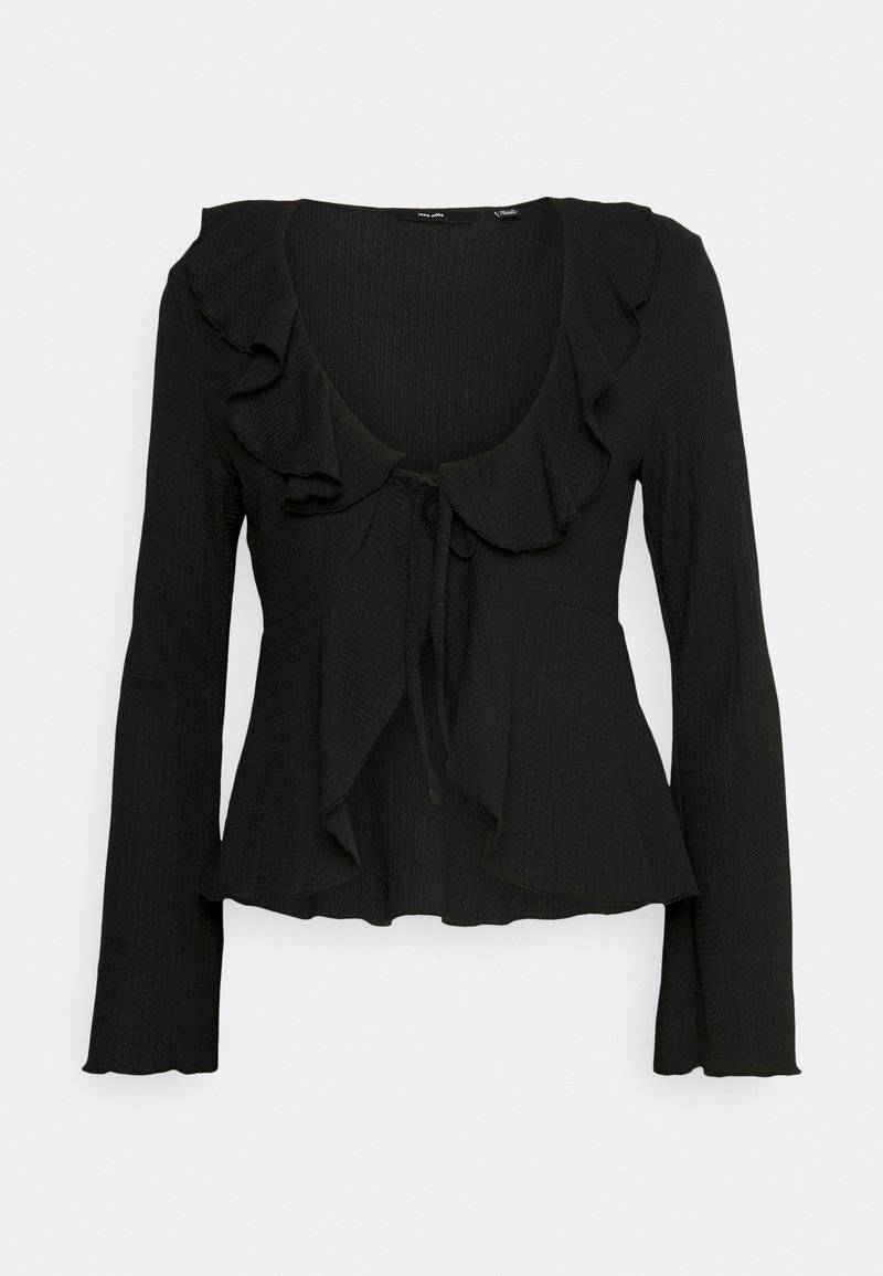 Blouse noire à manches longues avec col volanté et fermeture par nouer à l'avant, en tissu texturé avec un ourlet légèrement évasé.
