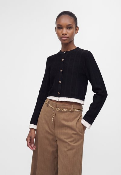 Femme portant un cardigan court noir boutonné sur un haut blanc avec un pantalon beige à fines rayures et une ceinture chaîne dorée, debout une main dans la poche.