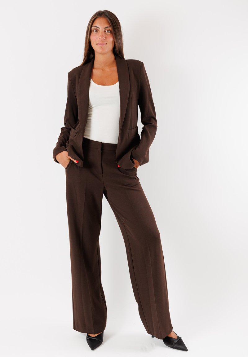 Completo marrone su misura con pantaloni a gamba larga e giacca aderente. Il completo presenta una texture liscia e due tasche laterali. Tacchi neri a punta.