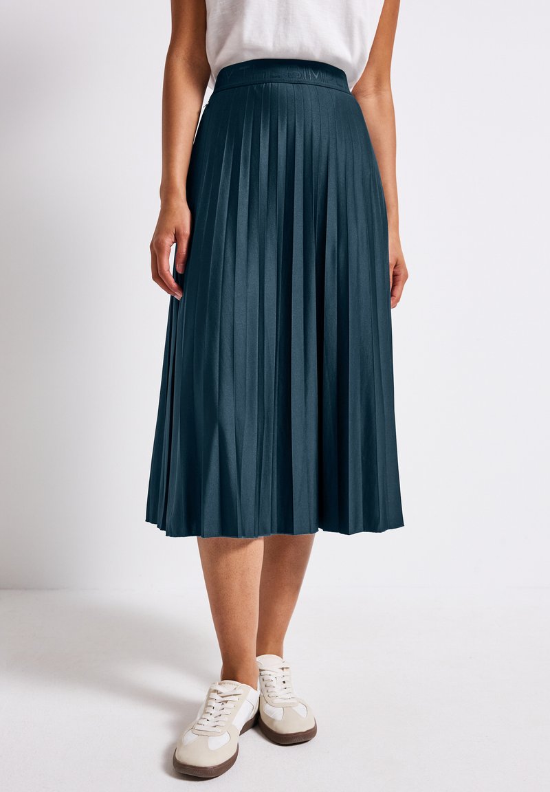 Jupe midi plissée bleu canard avec une texture lisse et une ceinture. Associée à des baskets blanches avec des accents beiges et sombres.