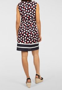 Femme portant une robe noire sans manches à pois blancs et rouges et des rayures horizontales blanches près de l'ourlet, assortie à des sandales compensées noires.