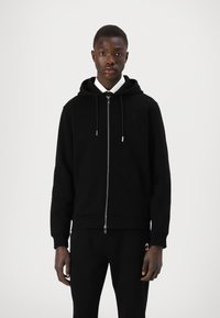 Sweatshirt preto com fecho, feito de tecido suave, com um fecho frontal, capuz ajustável com cordão e punhos canelados. Usado com calças pretas.