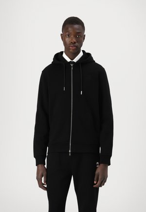 Felpa nera con zip realizzata in tessuto morbido, dotata di zip frontale, cappuccio regolabile con coulisse e polsini a coste. Abbinata a pantaloni neri.