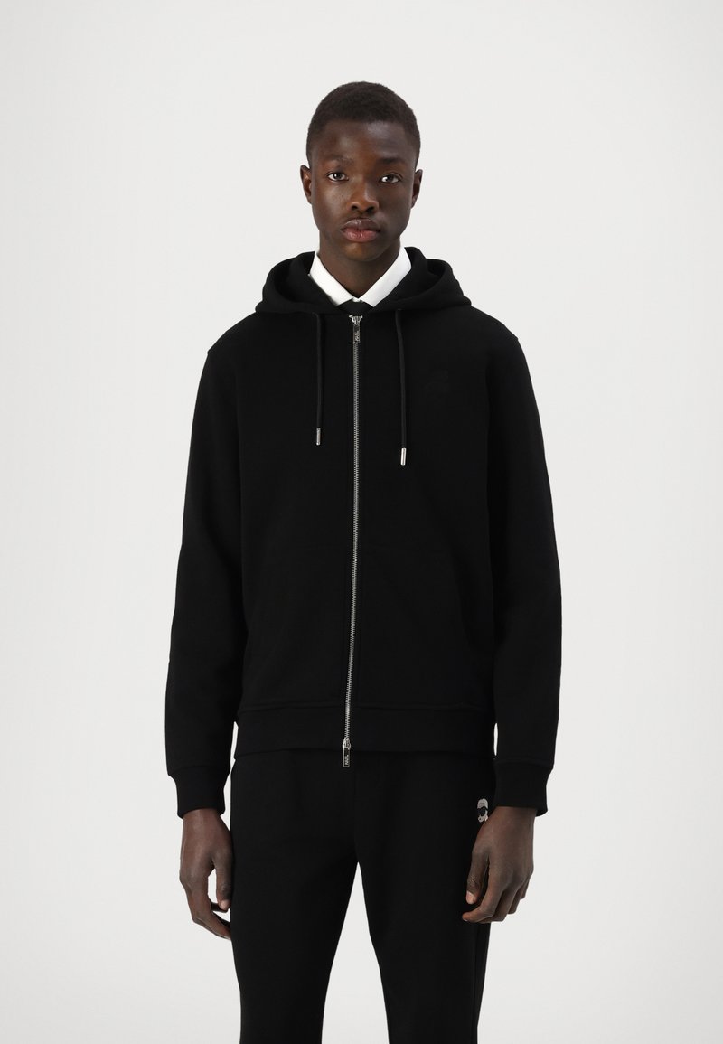 Sweatshirt preto com fecho, feito de tecido suave, com um fecho frontal, capuz ajustável com cordão e punhos canelados. Usado com calças pretas.