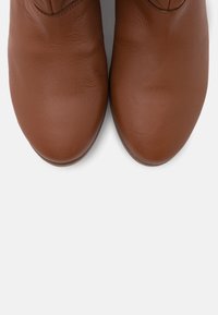 Bottes à cheville en cuir marron avec une texture lisse et un bout arrondi. Coutures minimales et aucun élément métallique visible, sur un fond blanc uni.