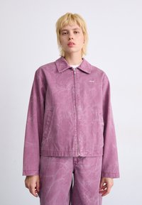 Rosa denimjakke med krage, frontglidelås og synlige sømmer. Har en slitt tekstur og en liten logo på venstre bryst.