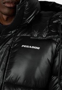 Muž v černé lesklé pufové bundě s vysokým límcem, na hrudi s bílým logem "PEGADOR".