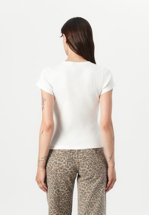 Femme aux longs cheveux foncés portant un haut blanc uni à manches courtes et un pantalon taille haute à imprimé léopard, debout de dos.