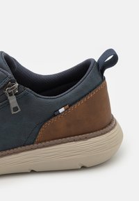 Zapato de caña baja en azul y marrón, hecho de cuero texturizado, con una cremallera, cuello acolchado y una suela de goma beige con estrías horizontales.