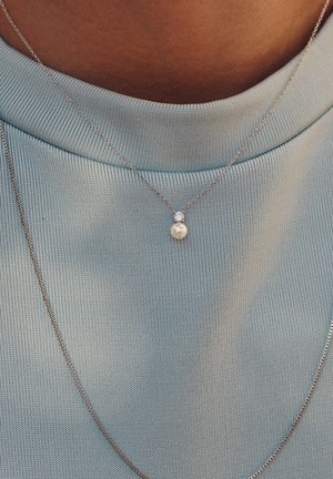 Silberne Halskette mit einem kleinen Perlenanhänger, der mit einem runden Diamanten akzentuiert ist, getragen über einem soften blauen Textiloberteil.
