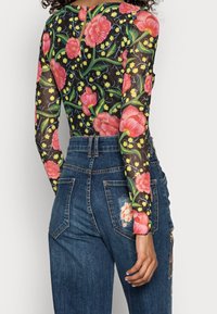 Långärmad, genomskinlig svart bodysuit med livfullt rosa och gult blommönster, kombinerad med högmidjade blå denimjeans med lappar.