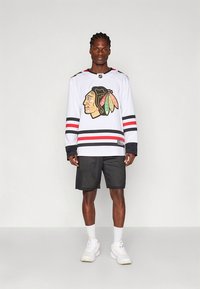 Vit hockeytröja med svarta och röda ränder och en färgstark laglogotyp, kombinerad med svarta meshningshorts och vita sneakers.