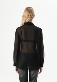 En blond kvinne som vender bort fra kameraet, har på seg en gjennomsiktig svart bluse med lange ermer som avslører en svart bh, sammen med teksturerte svarte bukser.