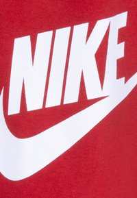 Sarkans audums ar drosmīgu baltu "NIKE" logo un izliekto balto swoosh dizainu, kas izceļ gludu tekstūru un izteiksmīgu grafikas stilu.