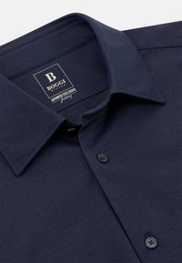 Temno modra srajca s gumbi iz teksturirane tkanine. Ima razprostranjen ovratnik in dve črni gumbe spredaj. Nalepka glasi "BOGGI Japonska ekskluziva."