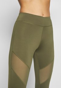 Olivgrön leggings med meshpaneler på sidorna, hög midja och mjuk textur. Sömnadsdetaljer längs sömmarna förbättrar designen.