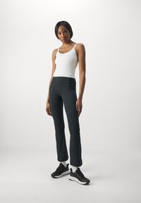 Sorte buksesnittutstyr med glat tekstur, parret med en hvid tætsiddende tanktop. Modellen har sorte atletiske sko med hvide såler.