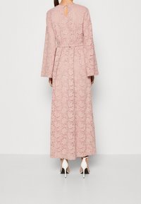 Długa różowa sukienka z koronki z długimi rękawami, wycięciem w kształcie łezki na plecach oraz paskiem w talii, w połączeniu z białymi szpilkami. Gładka faktura.