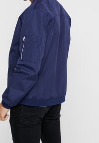 Námořnicky modrá bomber bunda z lehkého materiálu, s kapsou na zip na levém rukávu a elastickými manžetami v pase.