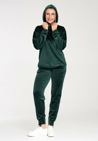 Grüner Velours-Trainingsanzug mit Kapuze, der eine lässige Passform und gerippte Bündchen aufweist. Kombiniert mit weißen Sneakers für einen Kontrast.