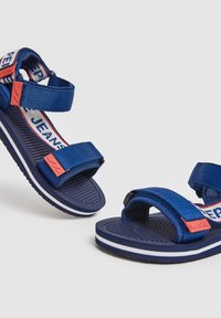 Pár modrých otevřených sandálů s nastavitelnými pásky, bílými a červenými akcenty, texturovanými podrážkami a látkovými detaily s logem.
