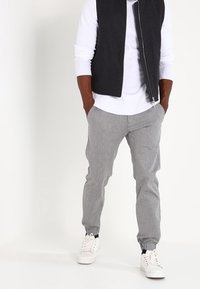 Gilet noir sans manches sur une chemise à manches longues blanche, associé à un pantalon gris avec revers et des baskets blanches. Tissu lisse, coupe décontractée.