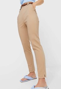 Beżowe spodnie o dopasowanym kroju z wysokim stanem, szlufkami na pasek i bocznymi rozcięciami na zakończeniach. Noszone z jasnoniebieskimi klapkami i pasującą górą.