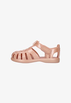 Sandale en plastique rose clair avec orteils fermés, multiples découpes latérales et bride réglable à la cheville sur fond blanc.