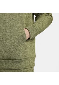 Main d’homme partiellement dans la poche avant d’un sweat-shirt texturé à manches longues, vert olive, sur fond blanc.
