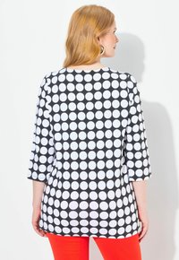 Schwarz-weiße Tupfen-Tunika mit Dreiviertelärmeln und rundem Ausschnitt, kombiniert mit leuchtend roten Hosen. Glatte Stofftextur.