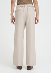 Pantalon beige clair à jambes larges avec une coupe décontractée, taille élastique et deux poches arrière, associé à des chaussures marron.