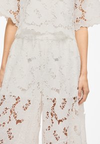 Weißes Spitzenzweiteiler-Outfit mit floralen Mustern, weiten Ärmeln und einem elastischen Taillenbund an der Hose, das ein zartes, luftiges Erscheinungsbild erzeugt.