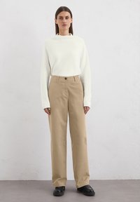 Pantalons larges beiges avec une fermeture à bouton, assortis à un pull blanc à manches longues et col rond. Tissu lisse et coupe décontractée.