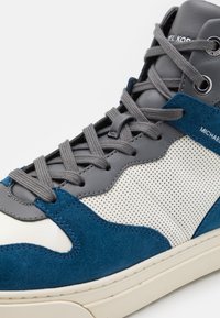 Vysoké tenisky s modrou semišovou a šedou koženou částí, s perforovanými bílými panely, šedými tkaničkami a logovými akcenty na límci.